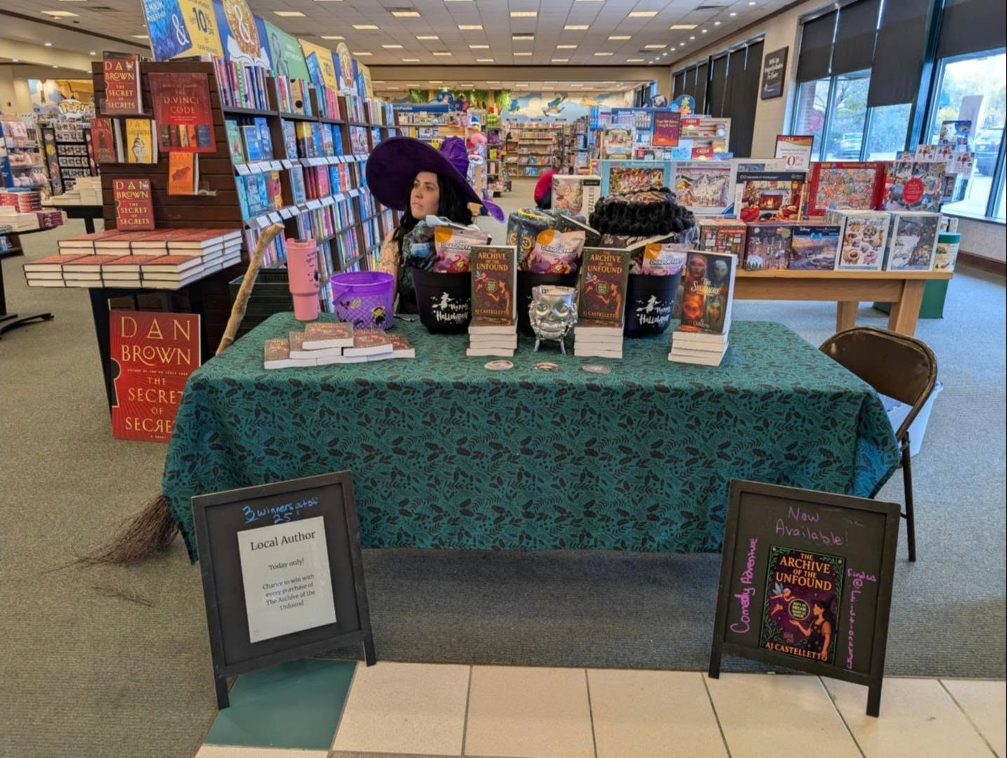 Aj Castelletto at a bookstore table in a purple witch hat, with stacks of The Archive of the Unfound and a sign that says Now Available